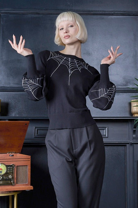 A model with a short blonde bob poses in a black knit sweater featuring white spiderweb embroidery on the chest and sleeves, paired with dark gray trousers, set against a dark paneled wall with a vintage radio in the foreground.