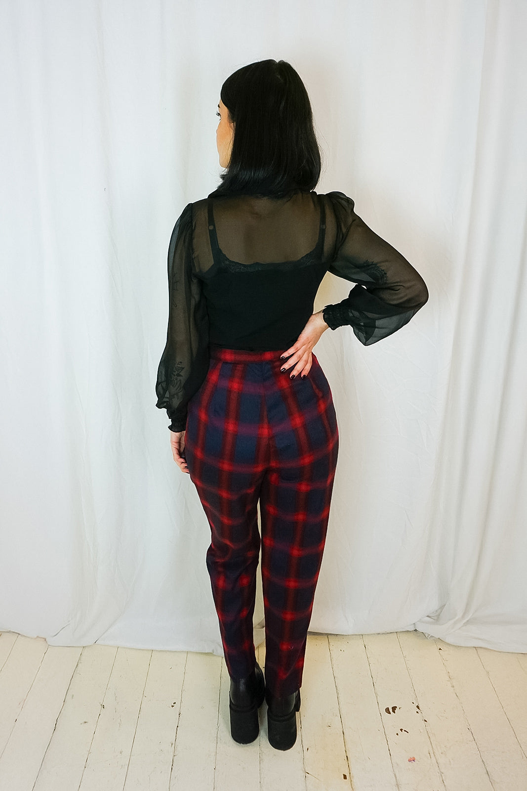 Person wearing a black sheer top and red plaid pants against a white curtain background